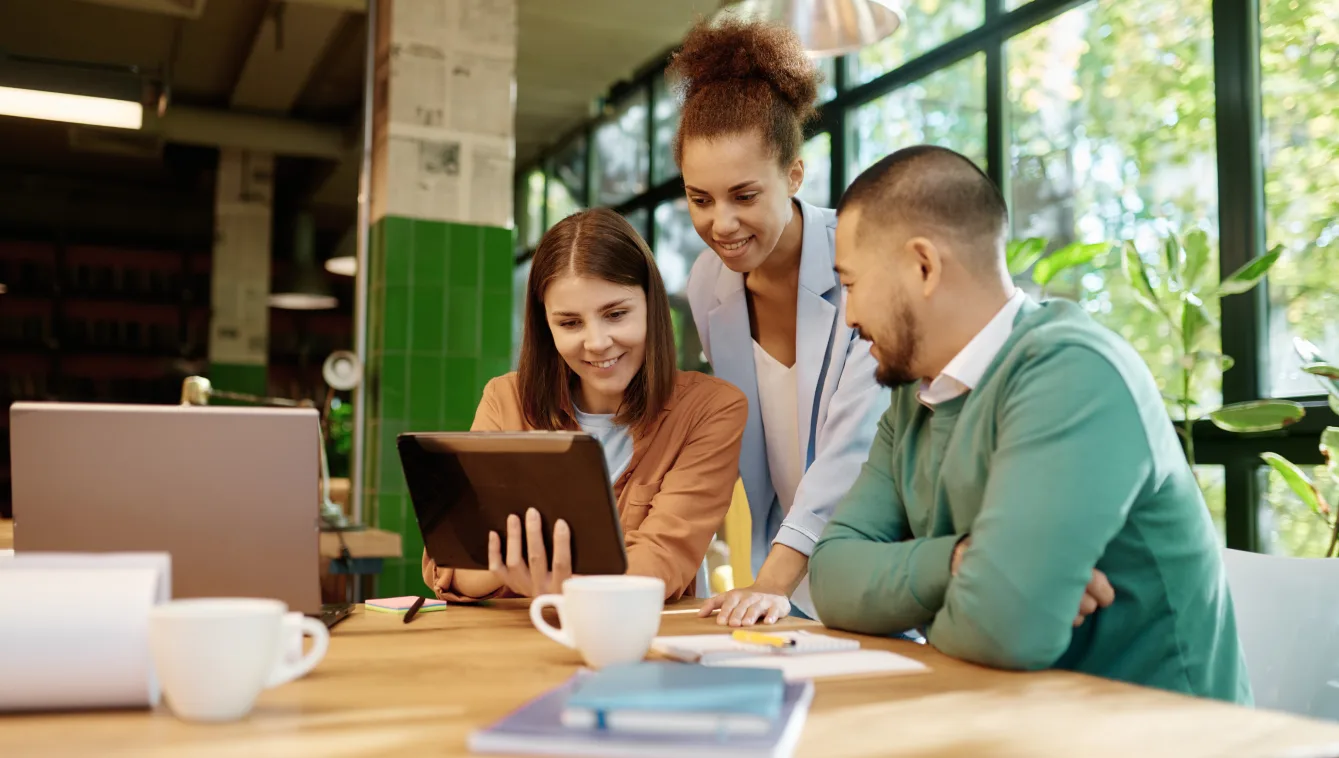 Three professionals are analyzing a tablet together at an office desk.